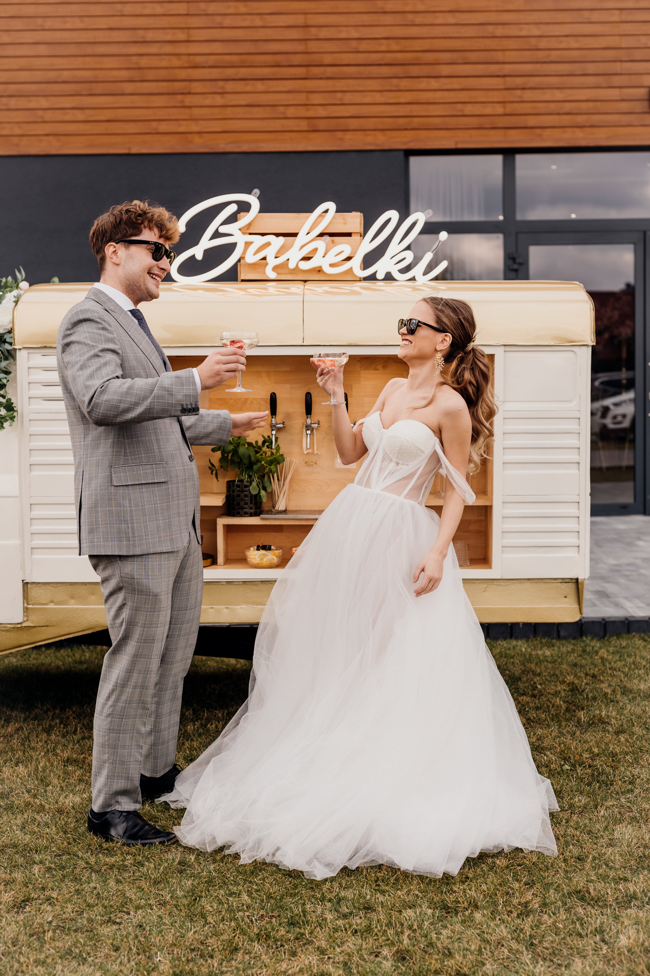 A groom and a bride posing with glasses of prosecco in the garden in front of a van