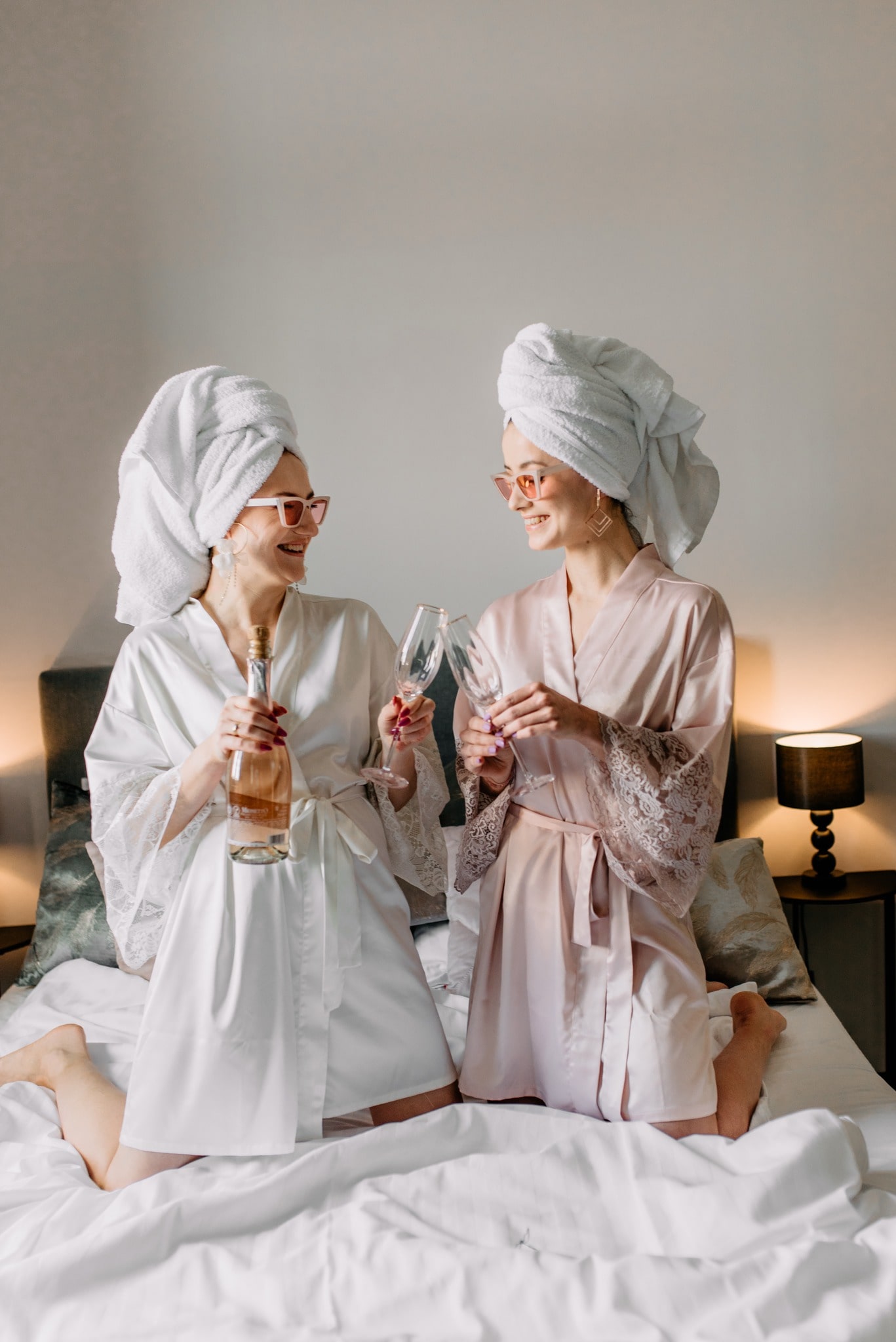 Two women in robes o the hotel bed posing with a prosecco and wine glasses